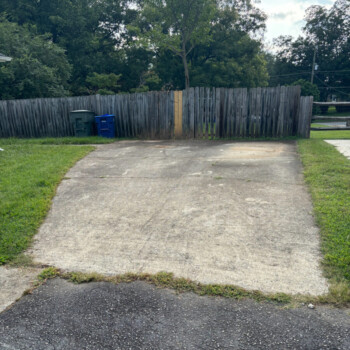 Empty concrete driveway leading to a wooden fence, with grass on both sides and trash bins near the fence.