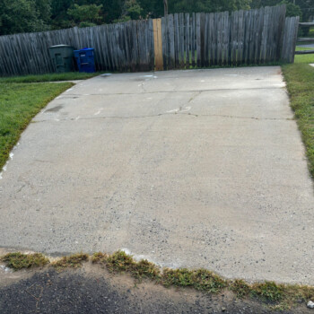 A cracked concrete driveway bordered by grass leads to a wooden fence with trash bins beside it.