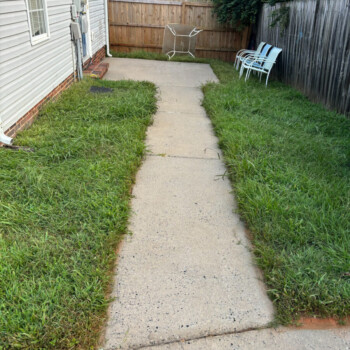 Concrete walkway bordered by overgrown grass, leading to a fenced area with chairs and an overturned clothes drying rack.
