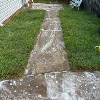 Concrete walkway with soap suds, surrounded by grass, leads to a fence; patio chairs and drying rack in background.