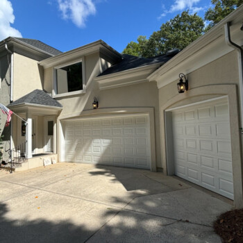 Two white garage doors on a beige house with outdoor wall lights, concrete driveway, and clear blue sky above.